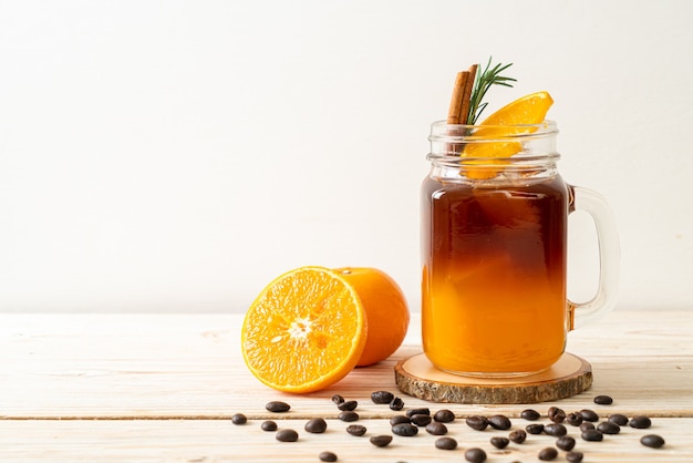 Un vaso de café negro americano helado y una capa de jugo de naranja y limón decorado con romero y canela en la mesa de madera