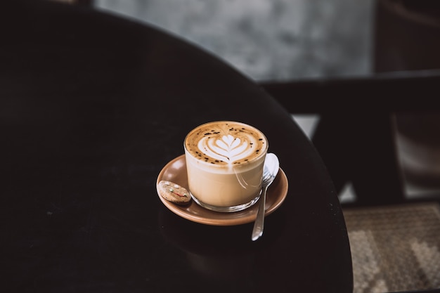 Vaso de capuchino con arte latte en platillo y con cuchara sobre la mesa negra. Bebida de desayuno