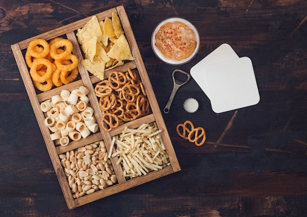 Foto vaso de cerveza artesanal y abridor con caja de snacks sobre fondo de madera. pretzel, palitos de papa salados, maní, aros de cebolla con nachos en caja vintage con abridores y tapetes de cerveza. vista superior