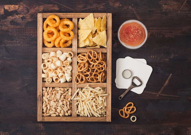 Vaso de cerveza artesanal y abridor con caja de snacks sobre fondo de madera. Pretzel, palitos de papa salados, maní, aros de cebolla con nachos en caja vintage con abridores y tapetes de cerveza.