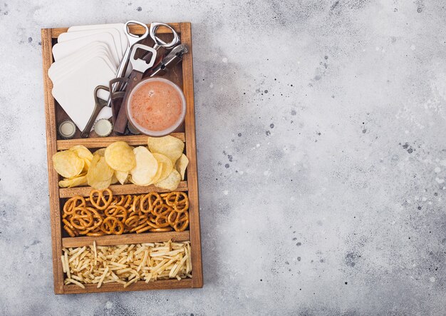 Vaso de cerveza artesanal en caja vintage de abrelatas y tapetes de cerveza en la mesa de la cocina ligera. Pretzel y patatas fritas y patata salada.