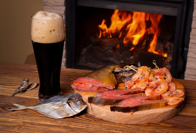 Vaso de cerveza oscura y una bandeja de pescado surtido sobre un fondo de una chimenea encendida