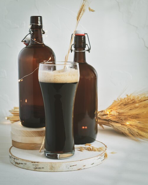 Foto vaso de cerveza oscura sin filtrar dos botellas marrones oscuras y un ramo de trigo sobre un fondo blanco