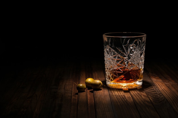 Vaso de cristal con whisky y aperitivos en una mesa de madera sobre un fondo negro