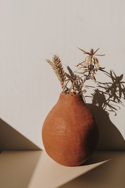 Vaso de flores de barro vermelho feito à mão com buquê de centeio de trigo seco em sombras de luz do sol em branco