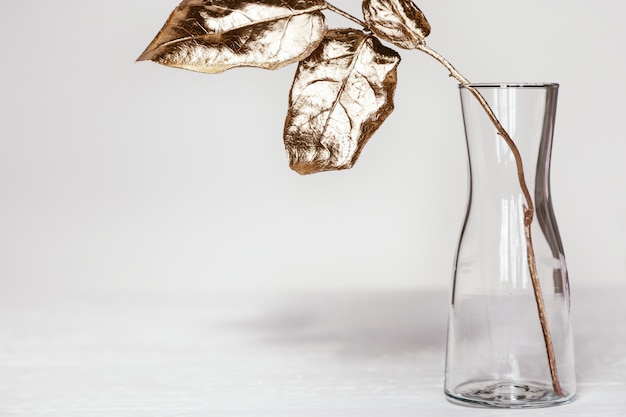 Vaso de vidro simples com galho de árvore e folhas coloridas ouro brilhantes na mesa branca. Planta de decoração para casa moderna.