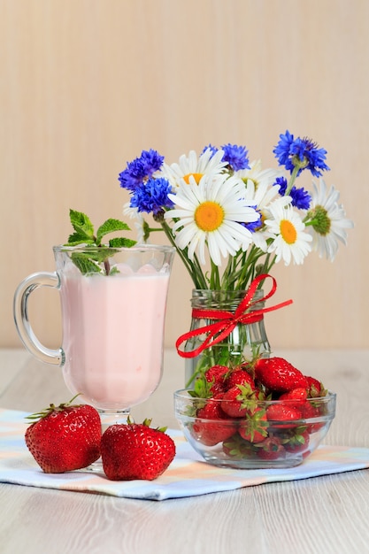 Vaso de delicioso yogur con menta y fresas frescas, manzanilla y acianos en un jarrón sobre una mesa de madera con una servilleta