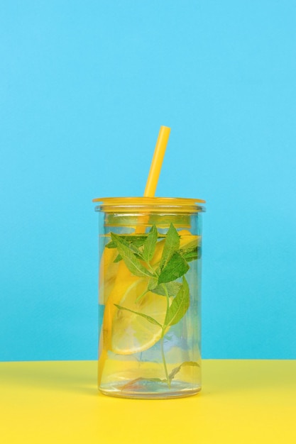 Un vaso grande con agua y limón y un tubo de cóctel sobre un fondo amarillo y azul.