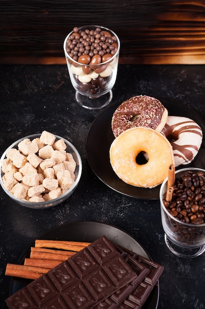 Vaso con granos de café junto a tabletas de chocolate, donas, azúcar morena y otro vaso con cacahuetes en chocolate sobre fondo de madera oscura.