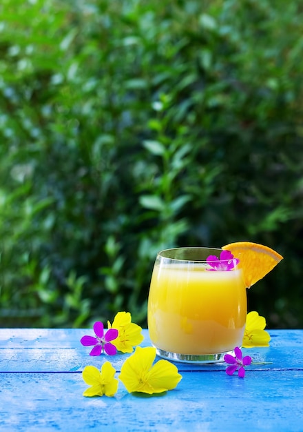 Vaso de jugo de naranja fresco adornado con una rodaja de naranja madura y flores en la mesa de madera azul