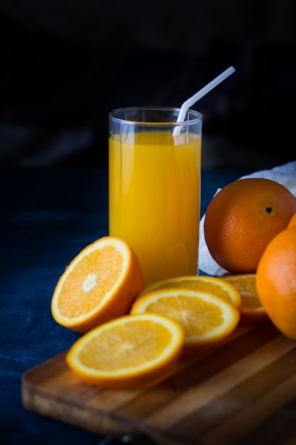 Un vaso de jugo de naranja fresco con un tubo, una mesa de cocina de madera, rodajas de naranja, naranjas, tela blanca sobre una superficie azul oscuro