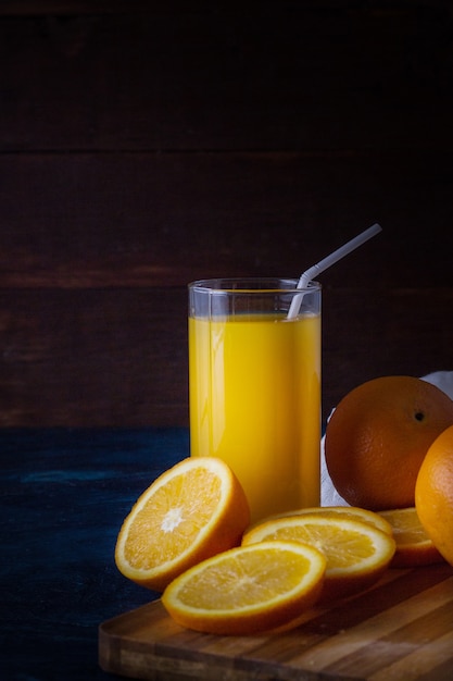 Un vaso de jugo de naranja fresco con un tubo, una mesa de cocina de madera, rodajas de naranja, naranjas, tela blanca sobre una superficie azul oscuro
