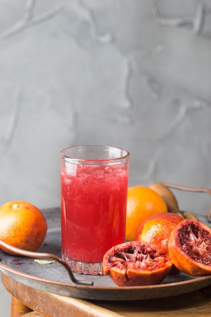 Vaso de jugo de naranja sanguina con hielo y frutas de naranja sobre fondo gris