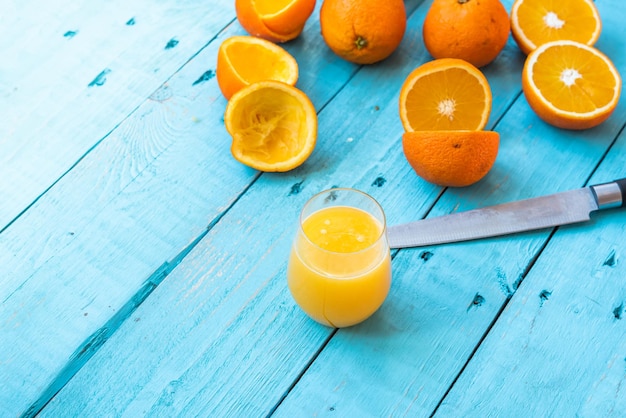 Vaso de jugo natural de varias naranjas orgánicas, algunas enteras, algunas exprimidas, para la dieta, sobre una mesa de madera azul. Desayuno saludable