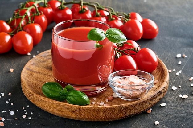 Vaso de jugo de tomate fresco, sal, albahaca y tomates en soporte de madera sobre fondo de madera vieja. Con espacio de copia.