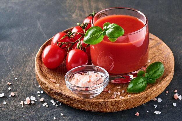 Vaso de jugo de tomate fresco, sal, albahaca y tomates en soporte de madera sobre fondo de madera vieja. Con espacio de copia.