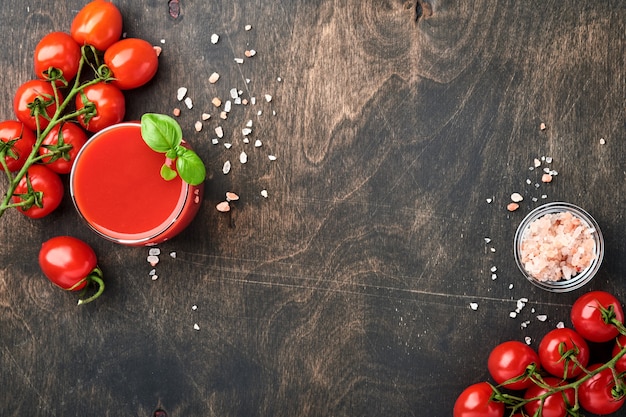 Vaso de jugo de tomate fresco, sal, albahaca y tomates en soporte de madera sobre fondo de madera vieja. Vista superior con espacio de copia.