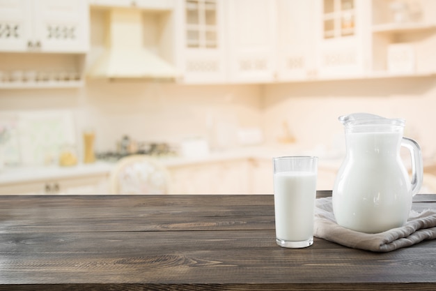 Vaso de leche fresca y jarra sobre mesa de madera con desenfoque de cocina como fondo.