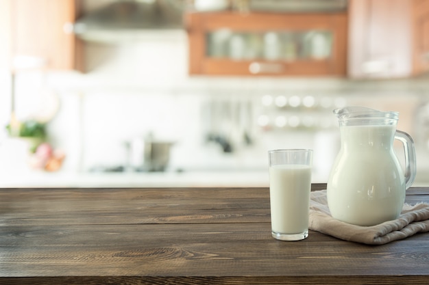 Vaso de leche fresca y jarra sobre mesa de madera con desenfoque de cocina como fondo.