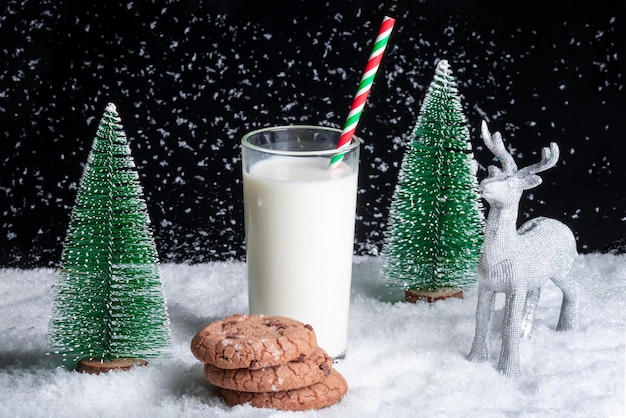 Un vaso de leche con pajita y galletas con trocitos de chocolate sobre un fondo nevado entre árboles de Navidad de juguete