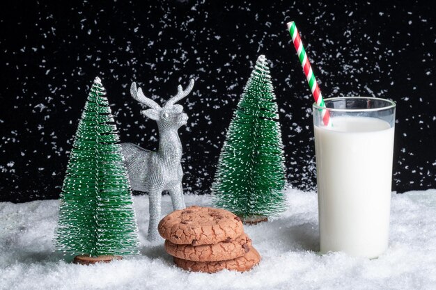 Un vaso de leche con pajita y galletas con trocitos de chocolate sobre un fondo nevado entre árboles de Navidad de juguete