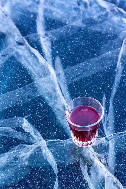 Un vaso con licor rojo se encuentra sobre el hielo de un lago con hermosas grietas. Alcohol en un vaso y hielo espolvoreado con nieve. Vertical.