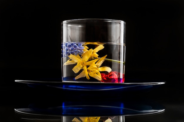 Un vaso de líquido transparente que contiene flores amarillas y azules y trozos rojos de hielo flota en un plato azul sobre una mesa de vidrio.