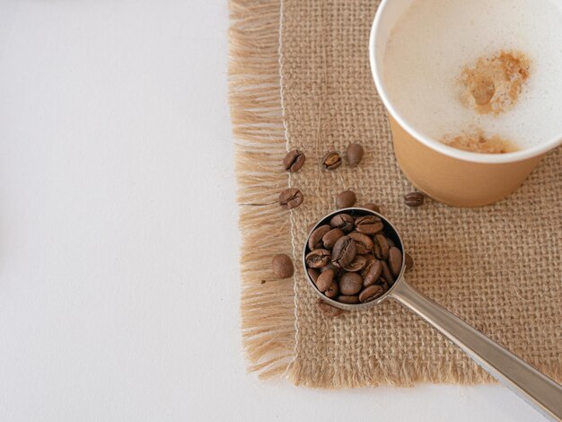 Un vaso de papel desechable con capuchino en una servilleta de lona, granos y café molido están esparcidos sobre la mesa y una cuchara para servir Vista desde arriba Espacio para copiar