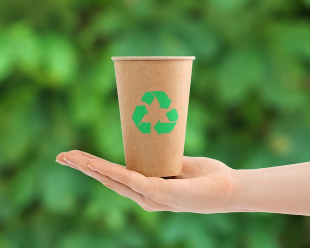 Vaso de papel desechable con signo de reciclaje en una mano femenina contra verdes borrosos