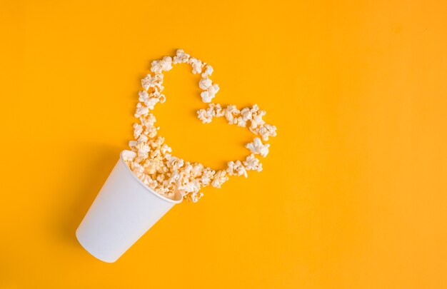 Vaso de papel con palomitas de maíz en forma de corazón sobre fondo amarillo. Banner endecha plana, vista superior. Ir al concepto de cine. Me gusta ver películas.