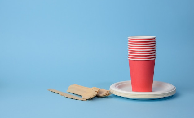 Vaso de papel rojo, platos blancos y tenedores de madera y cuchillos sobre una superficie azul. Concepto de rechazo de plástico, cero residuos, espacio de copia