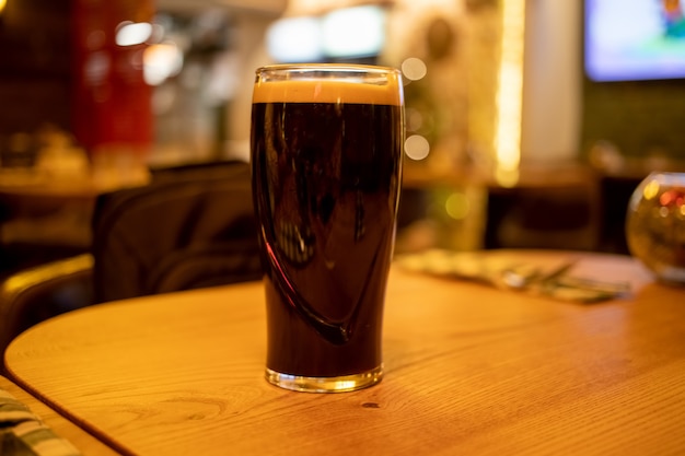 vaso de pinta de cerveza negra oscura con espuma se encuentra sobre una mesa de madera en un bar.