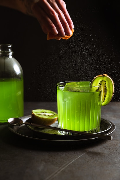 Un vaso bajo con una refrescante bebida verde y un cubito de hielo en su interior, adornado con kiwi, una botella de bebida verde, una cuchara de bar. Tiro de expresión de aceite de cítricos.