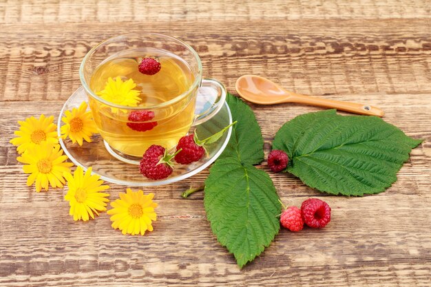 Vaso de té verde con flores de caléndula y frambuesas frescas sobre fondo de madera. Bebida saludable.