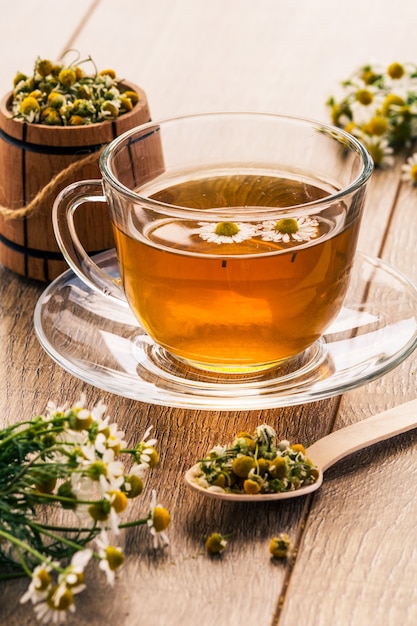 Vaso de té verde con flores de manzanilla blanca y barril de madera con flores secas de manzanilla en el fondo