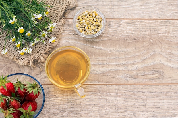 Vaso de té verde, flores de manzanilla, tazones de vidrio con flores secas de matricaria chamomilla y fresas frescas en el fondo de madera. Vista superior con espacio de copia.