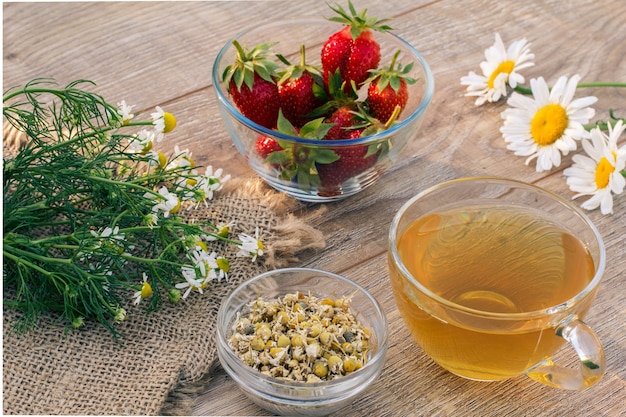 Vaso de té verde, flores de manzanilla, tazones de vidrio con flores secas de matricaria chamomilla y fresas frescas sobre las tablas de madera. Vista superior.