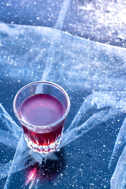 Un vaso de tintura se encuentra en el hielo del lago Baikal. Líquido rojo en un vaso que brilla al sol. Vista superior desde el lateral. Un poco de nieve sobre el hielo. Vertical.