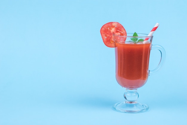 Vaso de vidrio con jugo de tomate, con tubo de cóctel y hojas de menta sobre fondo azul. El concepto de consumo de jugo de vegetales frescos.