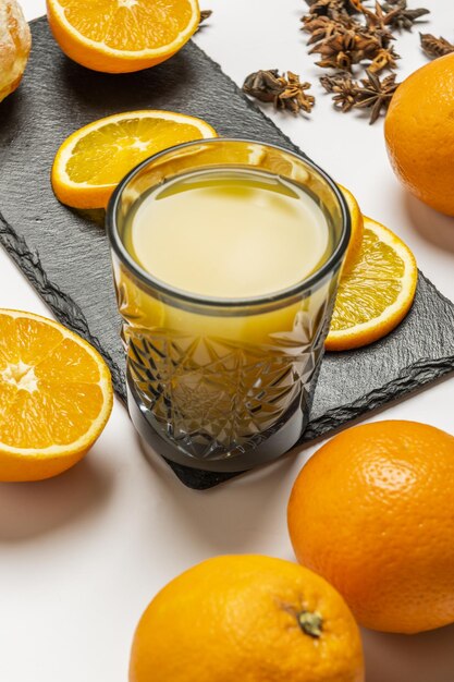 Un vaso de vidrio tallado lleno de jugo de naranja fresco rodeado de rodajas y naranjas enteras