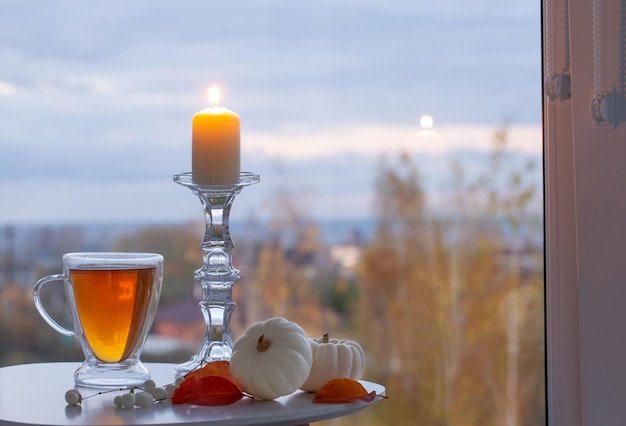 Vaso de vidrio con té y velas encendidas en la ventana de fondo con paisaje otoñal