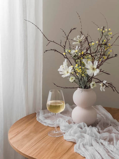 Un vaso de vino blanco y un jarrón con un ramo de primavera en una mesa redonda en la sala de estar
