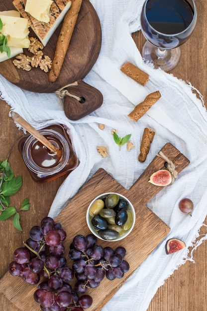Vaso de vino tinto, tabla de quesos, uvas, nueces, aceitunas, miel y palitos de pan en la mesa de madera rústica