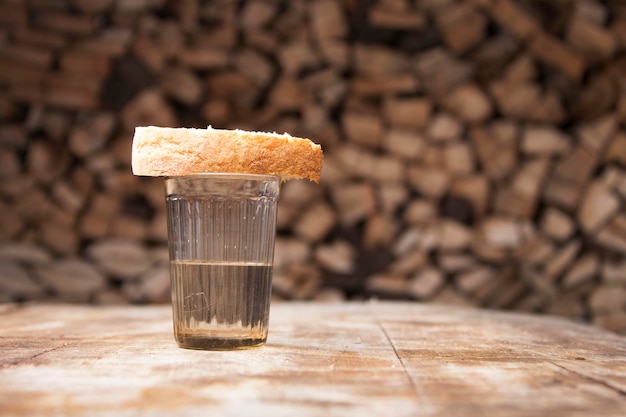 Un vaso de vodka y un trozo de pan sobre una mesa de madera y leña detrás