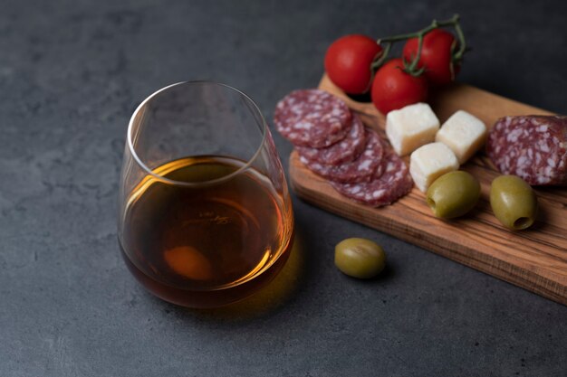 Vaso de whisky y bocadillos salchicha queso aceitunas y tomates sobre superficie de cemento gris oscuro