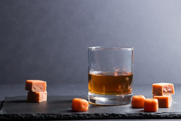 Vaso de whisky con caramelos de caramelo en un tablero de pizarra de piedra negra sobre superficie negra. Vista lateral, copia espacio.