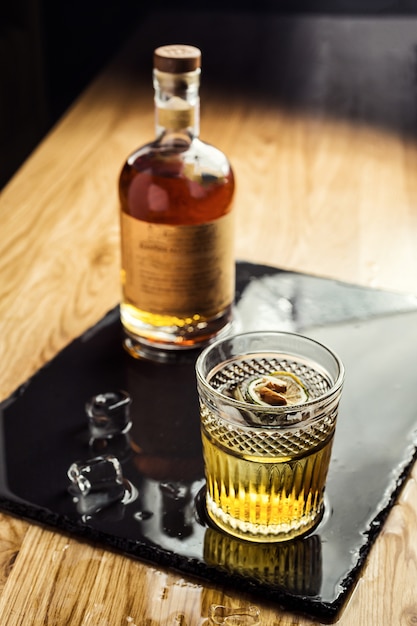 Vaso de whisky (coñac o brandy) con cubitos de hielo en la mesa oscura con una botella