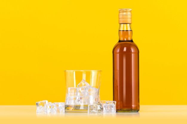 Vaso de whisky con cubitos de hielo y botella sobre fondo amarillo