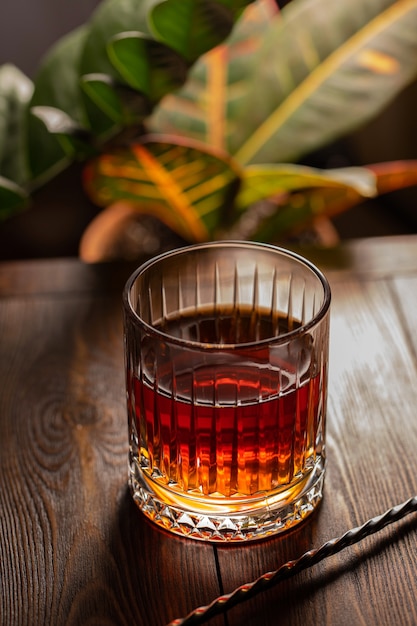 Vaso de whisky facetado con cuchara de cóctel sobre una mesa de madera en el fondo de plantas exóticas