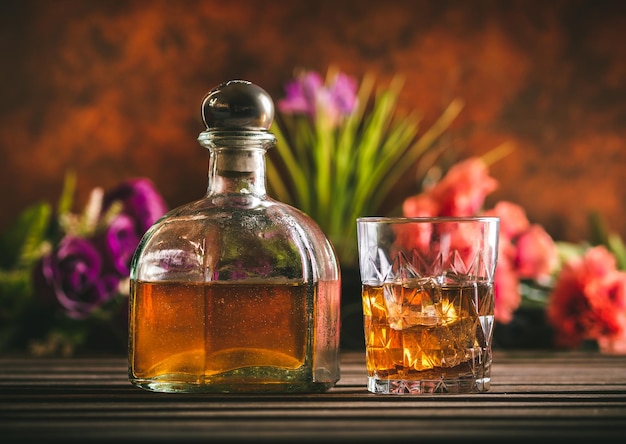 Vaso de whisky con hielo y botella con licor en una mesa de madera con algunas flores de color